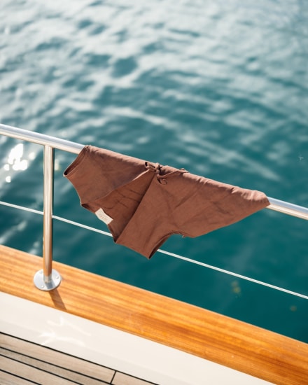 Prenda de lino marrón apoyada sobre la barandilla de un barco, con el mar azul de Menorca al fondo