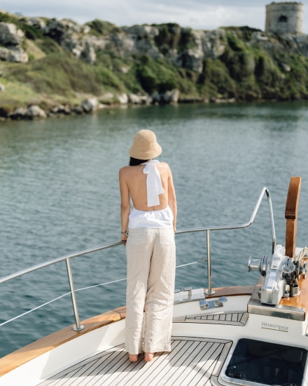 Mujer de espaldas apoyada en la proa de un barco frente a una cala de la costa de Menorca