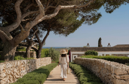 Huésped caminando por un sendero de piedra entre muros de marés en Torralbenc, con vistas al mar de Menorca